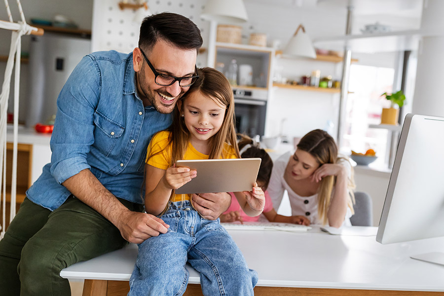 Client Center - A Happy Father is Embracing His Daughter While She Uses a Tablet and the Mother is Helping Their Other Daughter Behind Them at Home